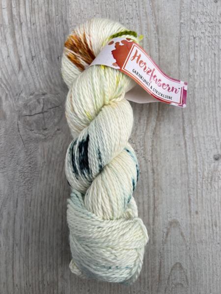 Ein Strang handgefärbtes Garn der Herzfaser No. 18, Grundfarbe naturweiß mit grünen, braunen und dunklen Sprenkeln. Liegt dekorativ auf hellem Holz.